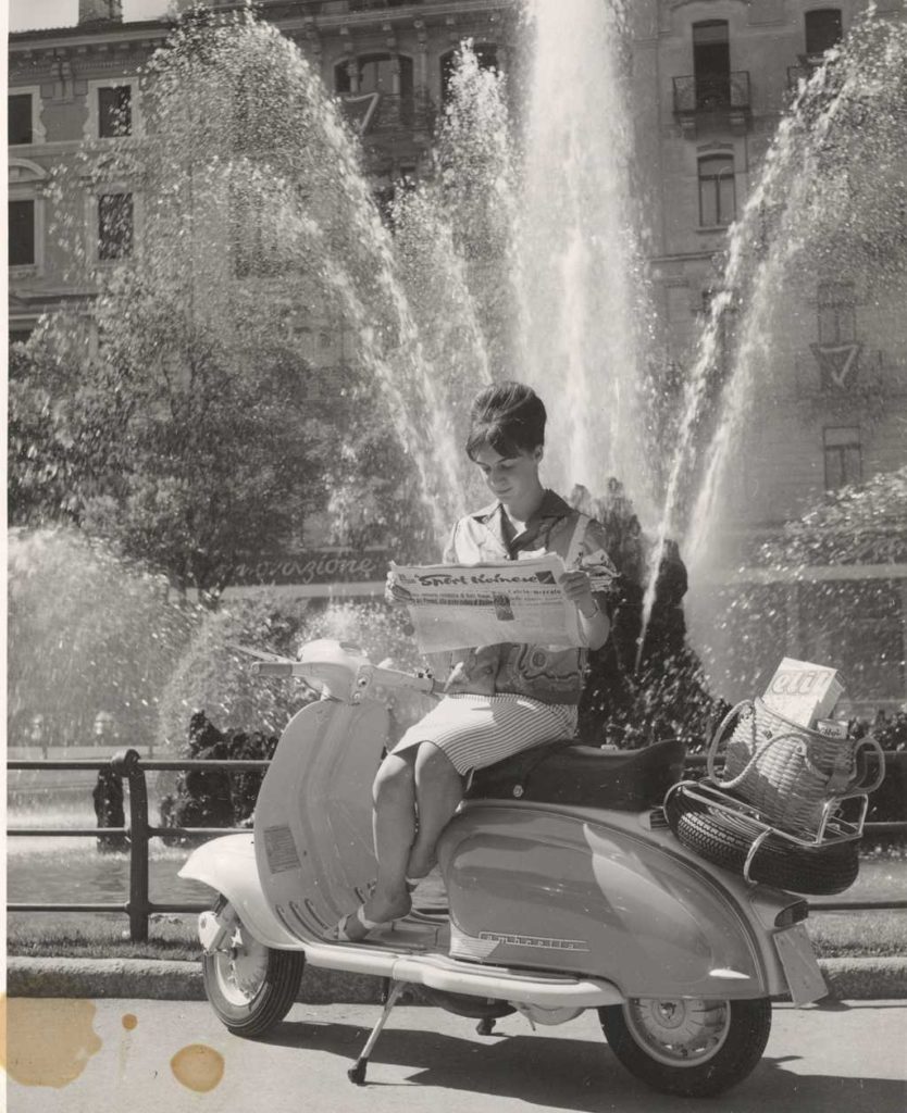 1960 lady reading paper on Lambretta scooter in frond of fountain